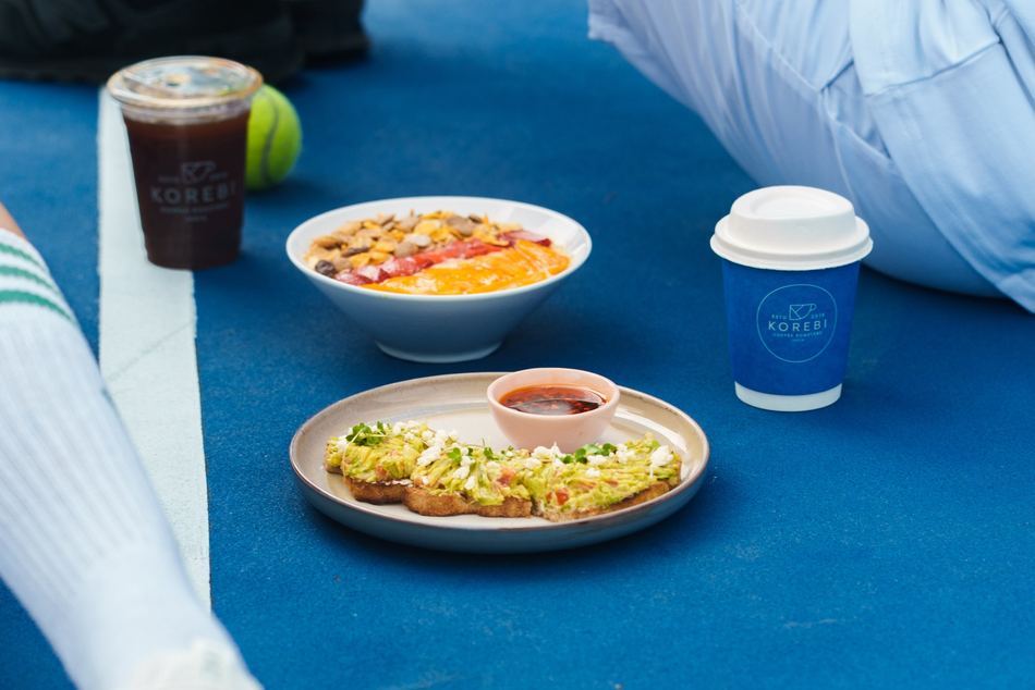 Hands holding Korebi coffee cups courtside at a padel court